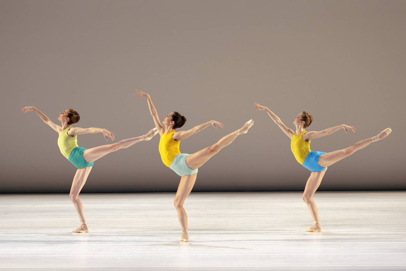 a trio of women bare-legged and bare arms wearing variations of yellow tank tops and blue green bottoms all in exquiste arabesques. Very high legs very upwardly reaching arms. Exuberant